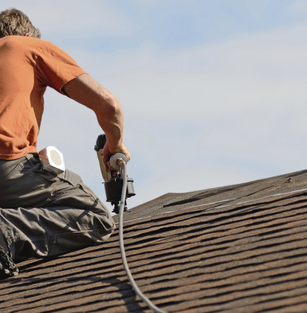Please create a brief 1 sentence description of the attached imageEditA shirtless roofer is using a pneumatic nail gun connected to an air hose to install or repair shingles on a sloped roof against a cloudy sky.